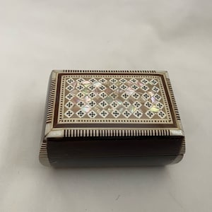 May include: A small, rectangular wooden box with a brown and white inlaid design. The top of the box is decorated with a geometric pattern of mother-of-pearl and black squares.