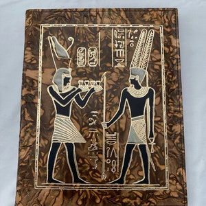 Könnte beinhalten: Ein braunes, ledergebundenes Tagebuch mit einem goldgeprägten Design von zwei Figuren im alten ägyptischen Stil. Die Figuren tragen aufwendige Kopfbedeckungen und halten einen Stab und ein Tablett mit Opfergaben. Der Hintergrund ist ein wirbelndes Muster aus Braun und Gold.