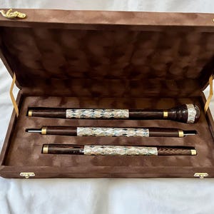 Könnte beinhalten: Ein braunes Samtetui enthält drei Schreibgeräte. Die Stifte und der Bleistift haben dunkle Holzschäfte mit silbernen und goldenen Akzenten. Der obere Stift hat ein abgerundetes Ende. Das Etui hat goldene Verschlüsse und Scharniere.