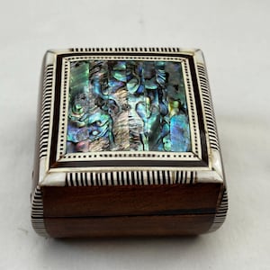 Puede incluir: Una pequeña caja de madera con una incrustación decorativa de concha de abulón iridiscente. La caja tiene un exterior de madera marrón oscuro con un borde rayado plateado y negro.