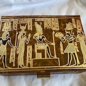 May include: A wooden box with a hinged lid and a drawer. The box is decorated with a gold and black design of Egyptian hieroglyphs and figures. The figures include a pharaoh sitting on a throne, a queen standing next to him, and other figures.