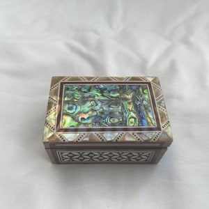 May include: Rectangular jewelry box with a decorative abalone shell inlay on the lid. The box has a brown wooden frame with geometric patterns and a rectangular abalone shell design in the center.