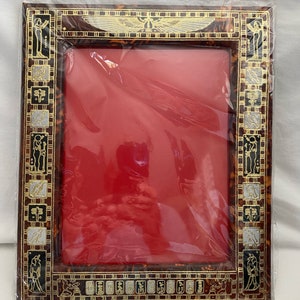 May include: A brown picture frame with a gold and silver design featuring Egyptian hieroglyphics and a winged sun disk. The frame has a red mount and is ready to display a photo or artwork.