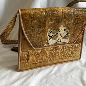 May include: A vintage brown leather shoulder bag with an embossed Egyptian design. The bag features a flap closure with a decorative emblem and a long strap. The bag's surface has a textured appearance, and the design includes hieroglyphs and figures.