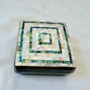 Pode incluir: Caixa quadrada com tampo em mosaico. O tampo é feito de pequenas telhas retangulares iridescentes em branco, verde e azul, dispostas em um padrão quadrado concêntrico. As laterais da caixa são marrom escuro.