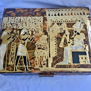 May include: A wooden box with an Egyptian theme. The box is decorated with gold and white paint and features a scene of Egyptian figures and hieroglyphics.