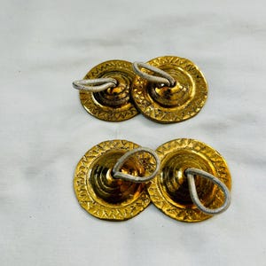 以下が含まれることがあります： 小さくて丸い金色のフィンガーシンバルが4つ。各シンバルはテクスチャ加工された表面と、保持するための白いコードのループが付いています。シンバルは2組に配置され、白い表面に置かれています。