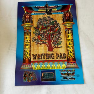May include: A blue and gold writing pad with an Egyptian theme. The front cover features a tree with birds, two columns with hieroglyphics, and a winged sun disk. The text "WRITING PAD" is printed in gold letters.