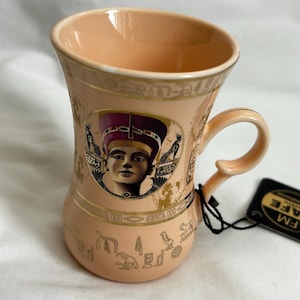May include: A peach-colored ceramic mug with a handle, featuring an image of an Egyptian pharaoh. The mug has gold-colored hieroglyphs and decorative bands. The interior of the mug is a lighter peach color. A black tag is attached to the handle.