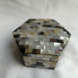 May include: A hexagonal shaped box made of mother of pearl tiles. The tiles are arranged in a brick pattern and are in shades of white, gray, and black.