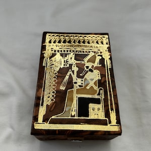 May include: A small wooden box with a brown and gold design. The box features a gold image of an Egyptian pharaoh sitting on a throne.