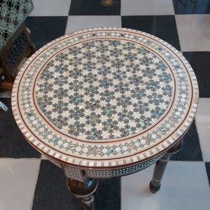 May include: A round, decorative table with a mosaic top featuring a geometric pattern of small, iridescent pieces. The table has a dark brown border and legs, and is set against a black and white checkered floor.