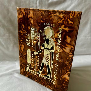 Könnte beinhalten: Ein braunes, ledergebundenes Notizbuch mit einem goldgeprägten ägyptischen Design. Das Cover zeigt eine Pharaonenfigur, Hieroglyphen und eine Sonnenscheibe. Das Notizbuch hat ein strukturiertes, marmoriertes Muster und einen genähten Rand.
