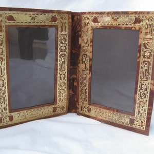 May include: A brown leather photo frame with gold accents and two openings for pictures. The frame has an Egyptian-inspired design with hieroglyphics.