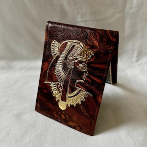 Könnte beinhalten: Eine braune Ledergeldbörse mit einem goldfarbenen, geprägten Design eines ägyptischen Pharaonenprofils. Die Geldbörse hat eine strukturierte Optik und ist leicht geöffnet, wodurch das Innere sichtbar wird.