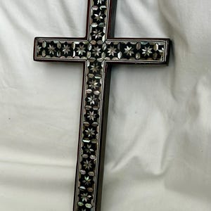May include: A wooden cross inlaid with mother-of-pearl in a geometric pattern. The cross is dark brown with a black background and white and iridescent mother-of-pearl accents.