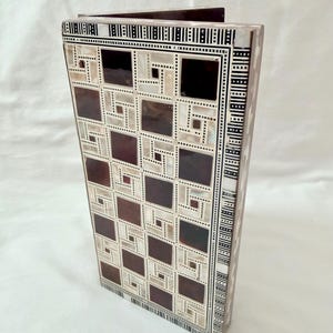 Puede incluir: Una caja decorativa de madera con un patrón geométrico. La caja presenta un diseño repetitivo de cuadrados en tonos marrones y blancos, con un borde de rectángulos blancos y negros. La caja está de pie sobre un fondo blanco.