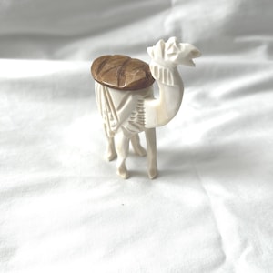 May include: A carved camel figurine with a cream-colored body and a brown saddle. The camel is standing on four legs, with its head raised and mouth open. The figurine is likely made of bone or a similar material.