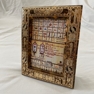 Pode incluir: Uma moldura dourada com um design intrincado que apresenta hieróglifos e símbolos egípcios. A moldura é decorada com um padrão de mármore marrom e dourado. A moldura contém uma folha impressa com uma tabela de hieróglifos e seus significados.