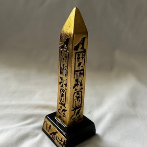 May include: A small gold obelisk with black hieroglyphics on all sides. The obelisk is on a black base with hieroglyphics.