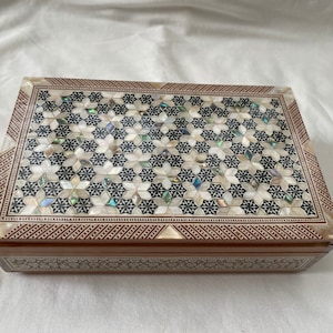 Puede incluir: Una caja de madera con un diseño decorativo de incrustaciones de nácar y madera negra. La caja tiene una tapa con bisagras y un patrón geométrico.