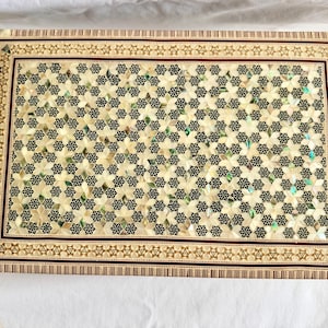 May include: A rectangular wooden box featuring an intricate geometric inlay pattern of cream-coloured and iridescent mother-of-pearl floral shapes set against dark wood. The border displays a repeating star motif and fine linear wood banding.