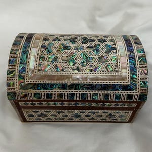 Puede incluir: Una caja de madera con tapa abovedada, finamente incrustada con nácar en un patrón geométrico. La caja es de color marrón oscuro y tiene un acabado brillante.