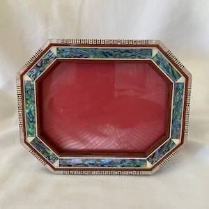 May include: An octagonal picture frame with a red interior. The frame is made of wood and features decorative inlays of iridescent abalone shell and a silver-coloured border. The frame is designed to hold a rectangular photo.