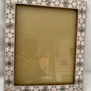 May include: A white picture frame with a decorative border made of iridescent mother of pearl tiles. The tiles are arranged in a geometric pattern with a red accent.