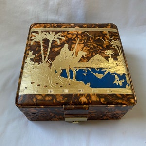 May include: A brown wooden box with a gold-colored design of a camel and a pyramid. The box has a hinged lid and a lock.