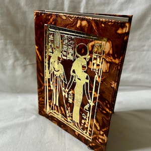 Könnte beinhalten: Ein braunes und goldenes Notizbuch mit ägyptischem Thema. Der Einband zeigt ein goldgeprägtes Design von zwei Figuren und Hieroglyphen. Das Notizbuch hat ein strukturiertes, marmoriertes Aussehen.