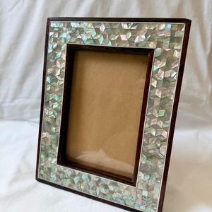 May include: A square photo frame with a dark brown wooden border. The frame is decorated with iridescent, geometric-shaped tiles. The interior of the frame is a light brown color, ready for a photo. The frame is set against a white background.