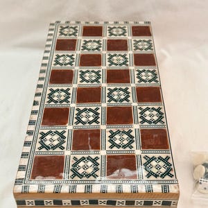 May include: A rectangular wooden backgammon board featuring a grid of alternating brown squares and white squares with intricate black and white geometric inlay patterns. The sides of the board are decorated with a repeating black and white border design.