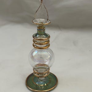 Può includere: Un flacone di vetro trasparente con un globo iridescente ornato di dorature e un tappo decorativo in vetro trasparente. Il flaconcino ha una base verde con finiture dorate.