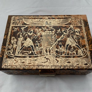May include: A wooden box with a brown marbled finish and a gold-colored design of Egyptian hieroglyphics and figures. The box has a hinged lid and a lock.