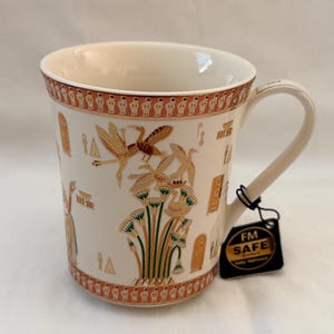 Könnte beinhalten: Ein cremefarbener Keramikbecher mit einem umlaufenden Muster aus kleinen Figuren am oberen und unteren Rand. Die Mitte zeigt goldene und grüne Illustrationen von Vögeln, Papyruspflanzen und Hieroglyphen. Ein schwarzes Etikett zeigt FM SAFE.