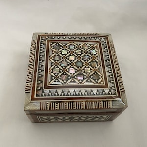 May include: A square wooden box with intricate inlaid designs. The box is made of wood and mother of pearl and has a brown, white, and black colour scheme.