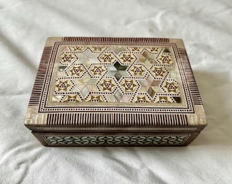 Puede incluir: Una pequeña caja de madera con un patrón geométrico rojo y blanco incrustado con nácar. La caja tiene una tapa con bisagras y está decorada con un diseño intrincado.