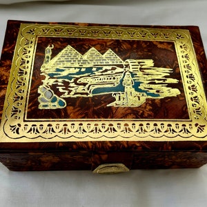 May include: A brown leather box with a gold-colored border and a scene of the pyramids of Egypt on the lid.
