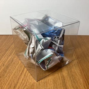 May include: A clear plastic cube box containing a crumpled piece of paper. The paper has a blue, white and red design.