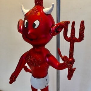 May include: Red glass Christmas ornament shaped like a devil with a white diaper and a red glitter pitchfork.