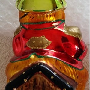 May include: A glass Christmas ornament shaped like a frog wearing a red and gold coat, sitting on a wooden treasure chest. The frog is wearing a green hat and has a gold trim.