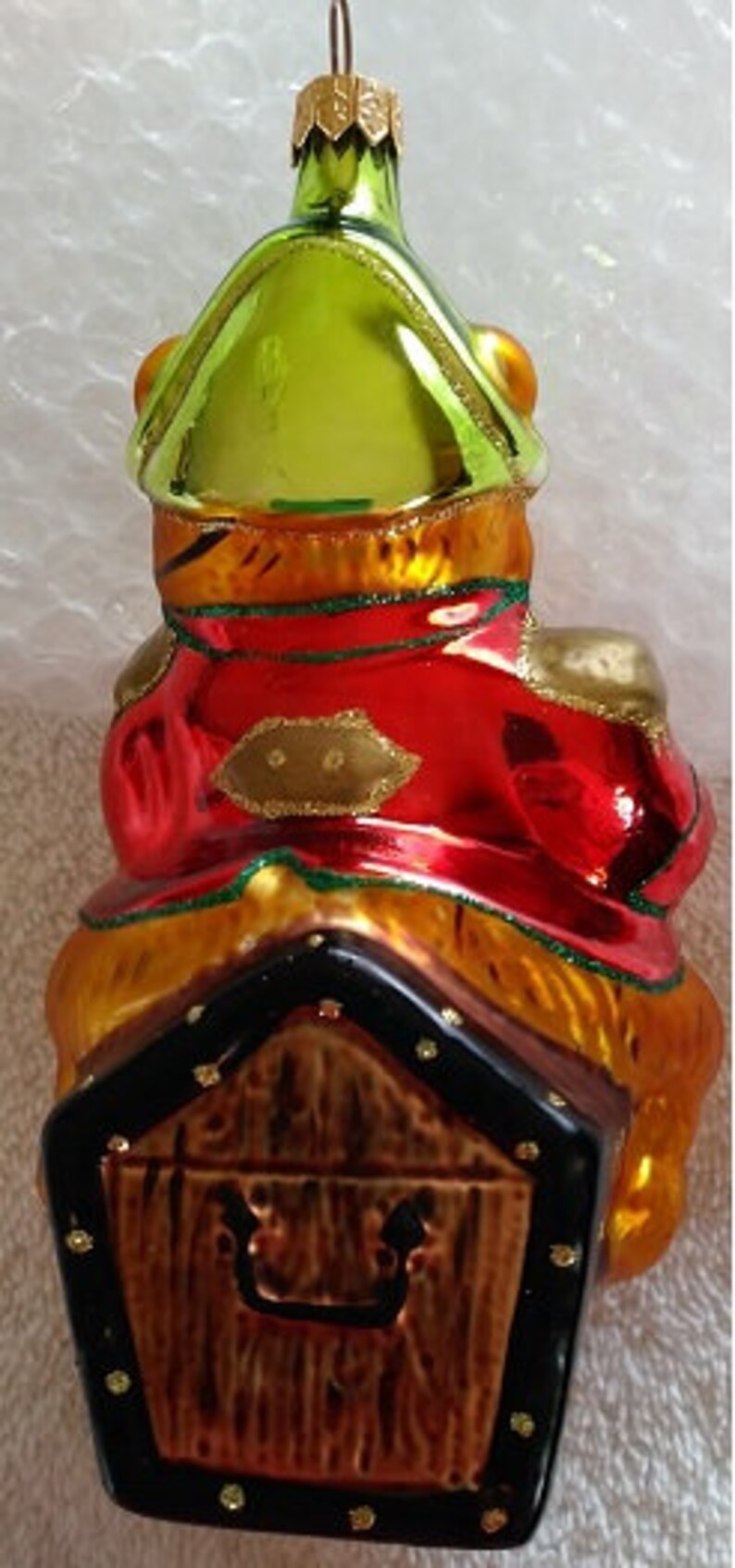 May include: A glass Christmas ornament shaped like a frog wearing a red and gold coat, sitting on a wooden treasure chest. The frog is wearing a green hat and has a gold trim.