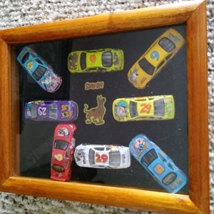 May include: A framed display of eight miniature toy race cars, featuring a Scooby Doo theme. The cars are yellow, blue, red, purple, and black with various numbers and cartoon characters. The frame is made of wood and has a glass front.