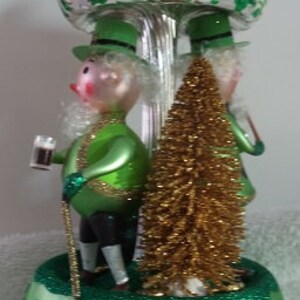 Puede incluir: Un adorno navideño de vidrio verde con dos duendes que sostienen una pinta de cerveza y un árbol. El adorno está decorado con purpurina y tiene una parte superior plateada.