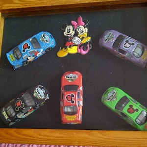 以下が含まれることがあります： ミッキーマウスとミニーマウスのキャラクターが描かれたミニチュアレーシングカー6台が額装されたディスプレイ。車は青、紫、黒、赤、緑など様々な色で塗装されています。各車にはミッキーマウス、ミニーマウス、ドナルドダック、グーフィー、プルートなど、異なるキャラクターが描かれています。フレームは木製で、背景は黒です。