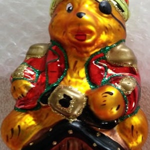 May include: A glass Christmas ornament shaped like a bear wearing a green pirate hat and an eye patch. The bear is holding a treasure chest and sitting on top of a small wooden house.