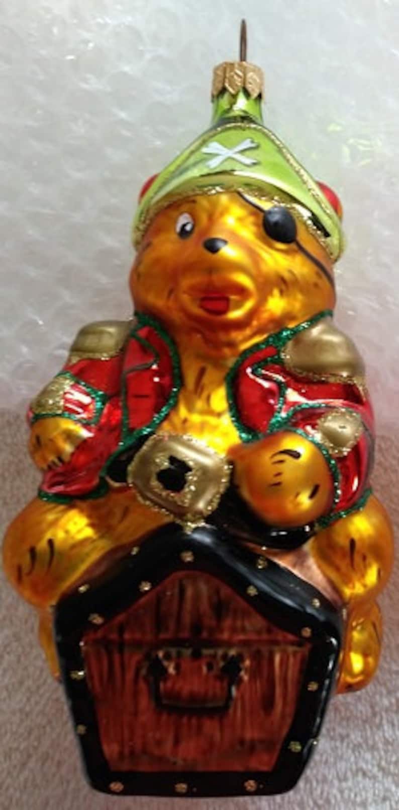 May include: A glass Christmas ornament shaped like a bear wearing a green pirate hat and an eye patch. The bear is holding a treasure chest and sitting on top of a small wooden house.