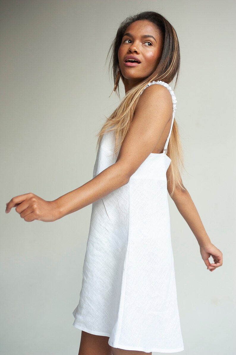 May include: A woman wearing a white sleeveless dress with a ruffled neckline. She is standing in front of a white background and looking to the left.