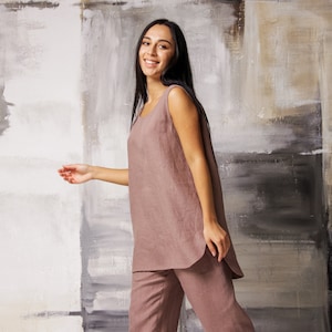 Op de afbeelding: Een vrouw draagt een loszittende, mouwloze linnen jumpsuit in een stoffige roze kleur. De jumpsuit heeft een rechte pijp en een licht afgeronde zoom. De vrouw staat voor een getextureerde muur met een neutrale kleurenpalet.